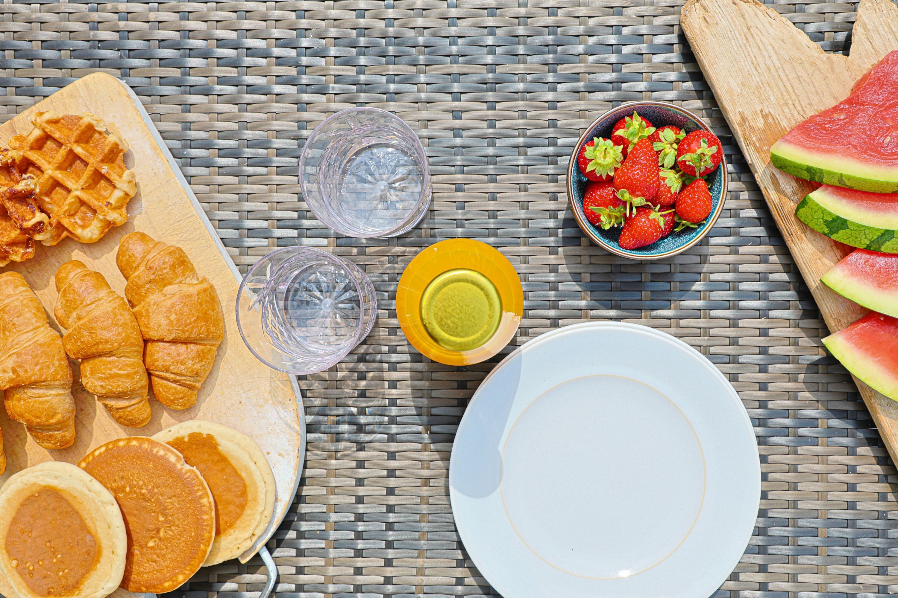 A breakfast setting with waffles, croissants, strawberries, watermelon, and glasses of water on a woven table.