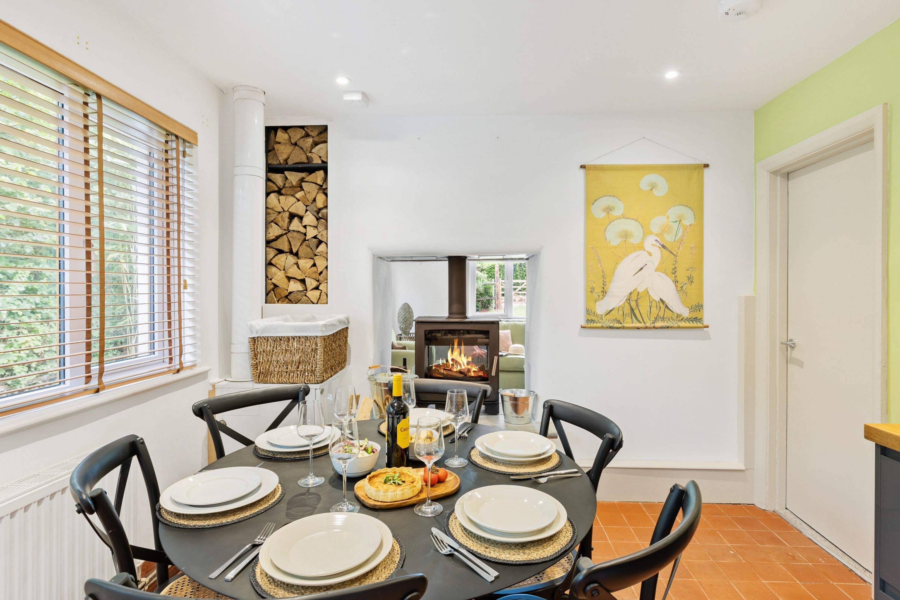 Un comedor con una mesa redonda dispuesta para cuatro personas, rodeada de sillas negras. Se ve una chimenea al fondo, y un cuadro amarillo cuelga en la pared.