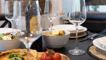 A dining table with a bottle of champagne, two empty wine glasses, a bowl of food, a plate with a pie, and two tomatoes on a wooden board.