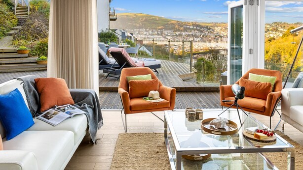A modern living room with a glass coffee table, two orange armchairs, a white sofa, and a view of a cityscape through large windows. - Homefield in Dartmouth (Dartmouth)