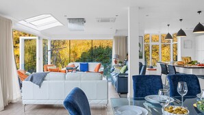 A modern living room with a sofa, dining table, and chairs. There is a view of trees through large windows. - Homefield in Dartmouth (Dartmouth)