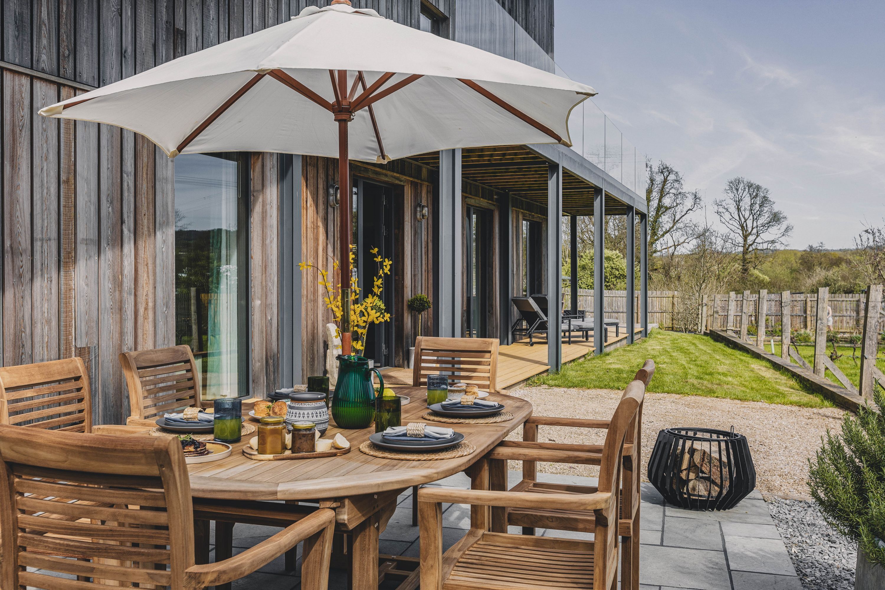 Ein Holztisch mit Stühlen, ein großer Sonnenschirm und ein Blumenkrug.
