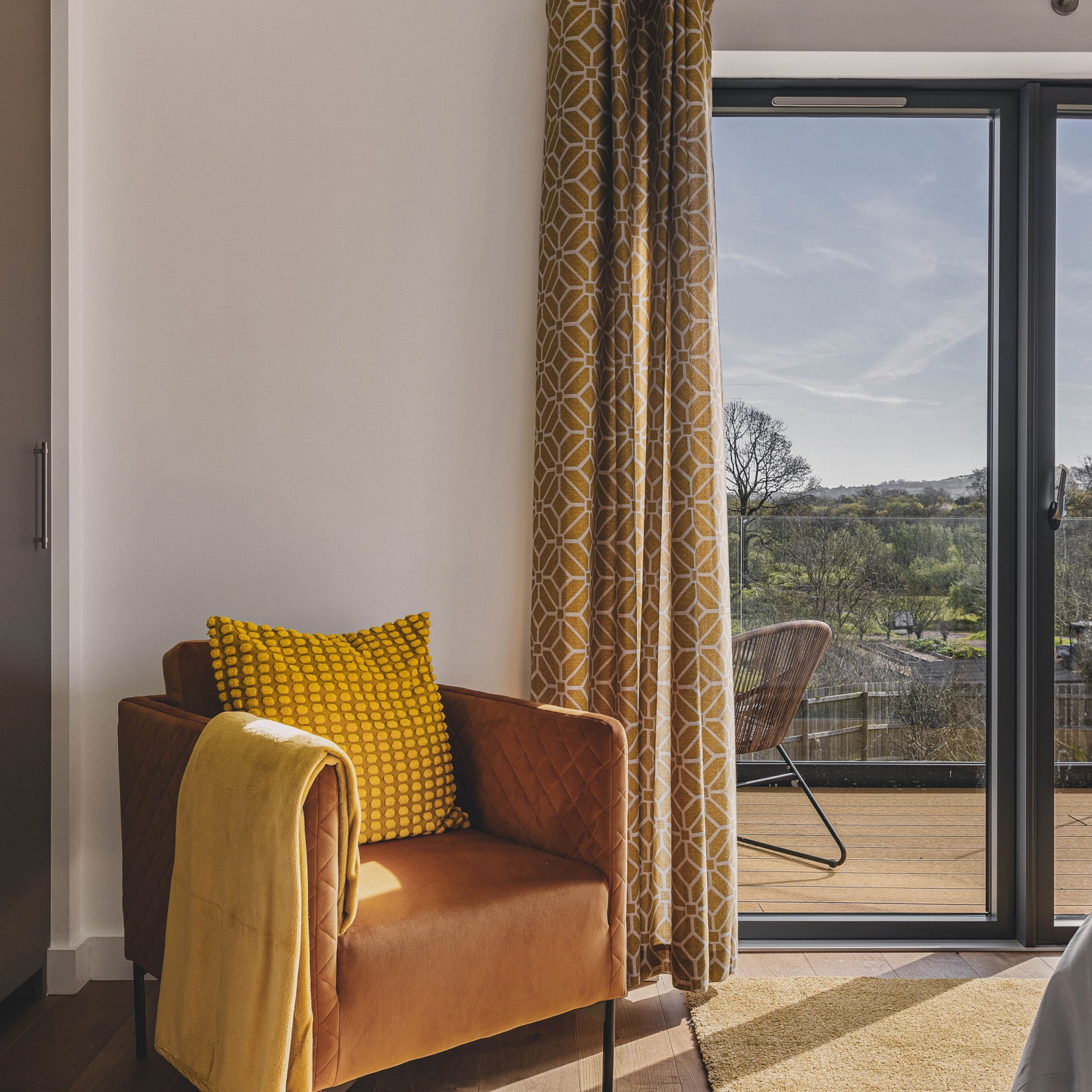 A modern living room with a brown sofa, yellow patterned pillows, and a large window with a view of a landscape.