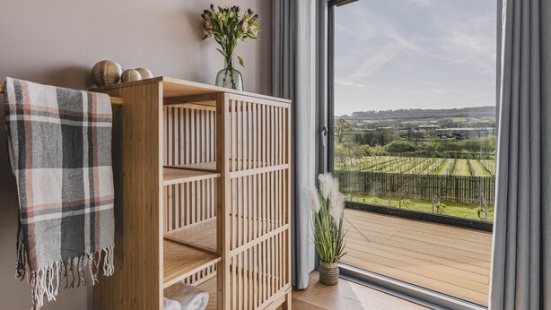 A room with a wooden shelf, a plaid towel, a vase with flowers, and a view of a vineyard through a large window.