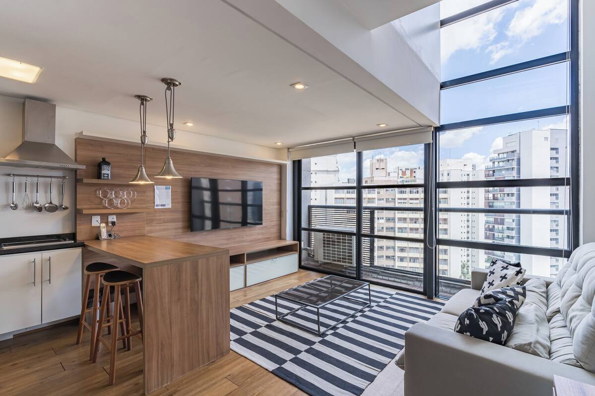 A modern kitchen with a wooden island, bar stools, and a large window offering a city view.
