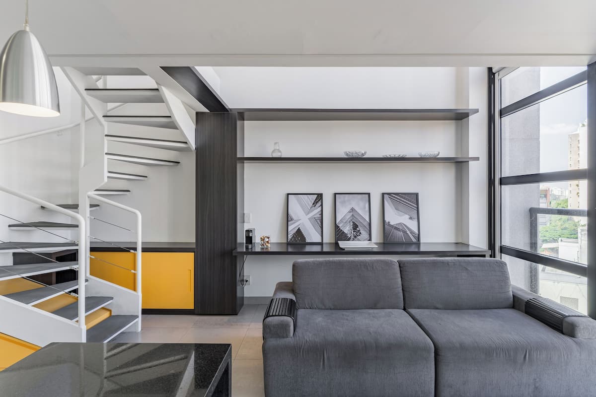 A modern living room with a grey sofa, a white spiral staircase, and a large window offering a city view.