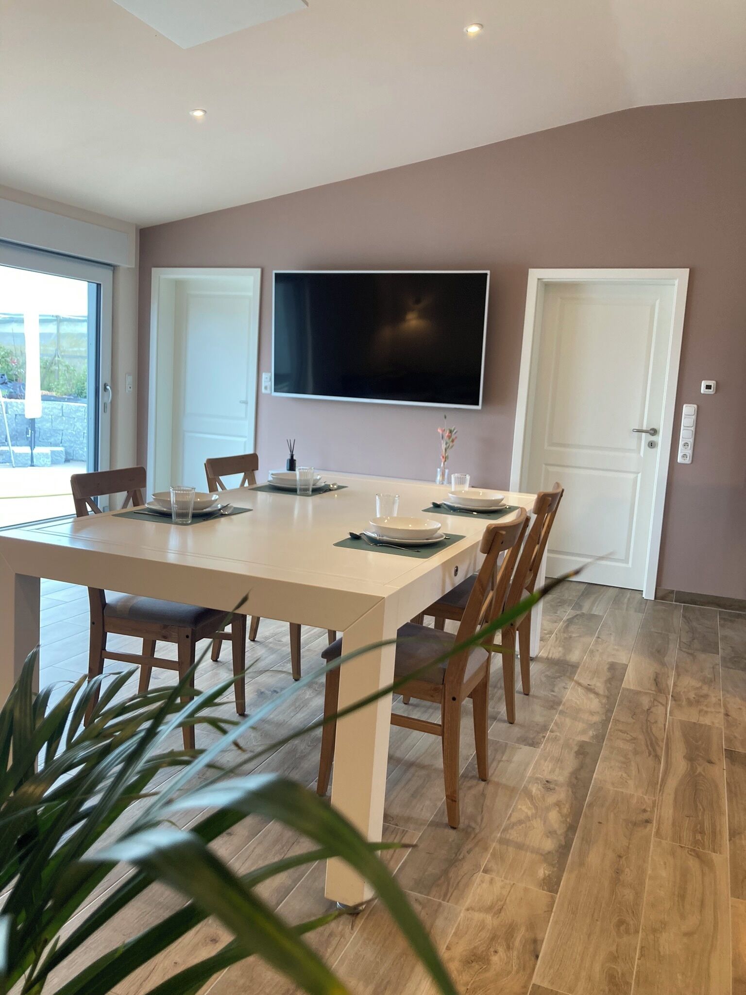 A modern dining area with a large table, chairs, and a flat-screen TV mounted on the wall.