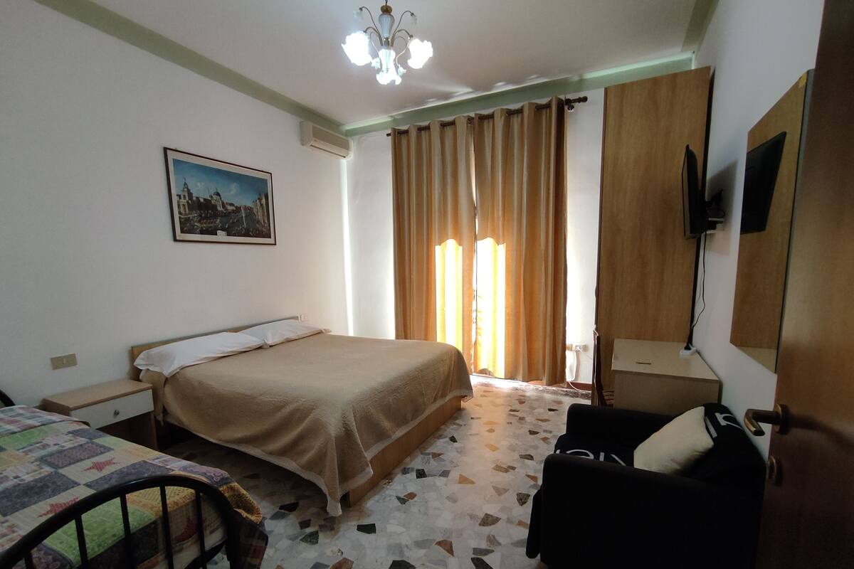 Une chambre à coucher comprenant un lit, une chaise, un bureau, un téléviseur et une fenêtre avec des rideaux.