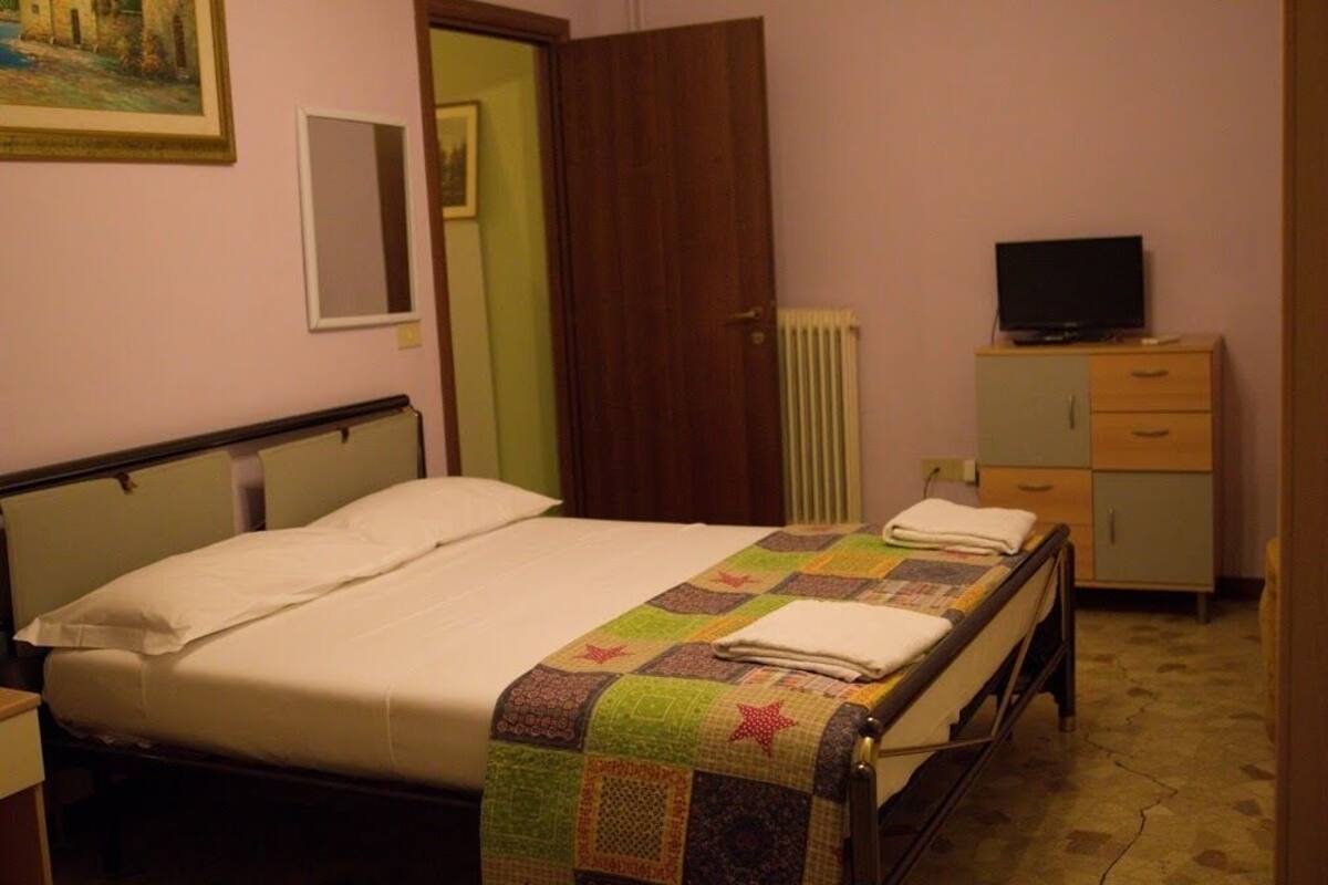 Une chambre avec un lit simple, une tête de lit en bois, une courtepointe, une table de chevet, un téléviseur posé sur un meuble et une porte donnant sur une autre pièce.