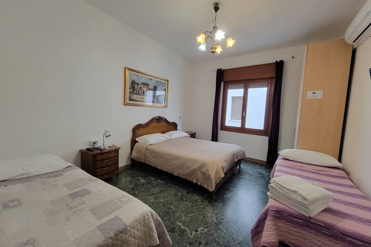 Une chambre à coucher comprenant un lit, une table de chevet, une commode, une fenêtre avec des rideaux, un tableau au mur et un appareil de climatisation.