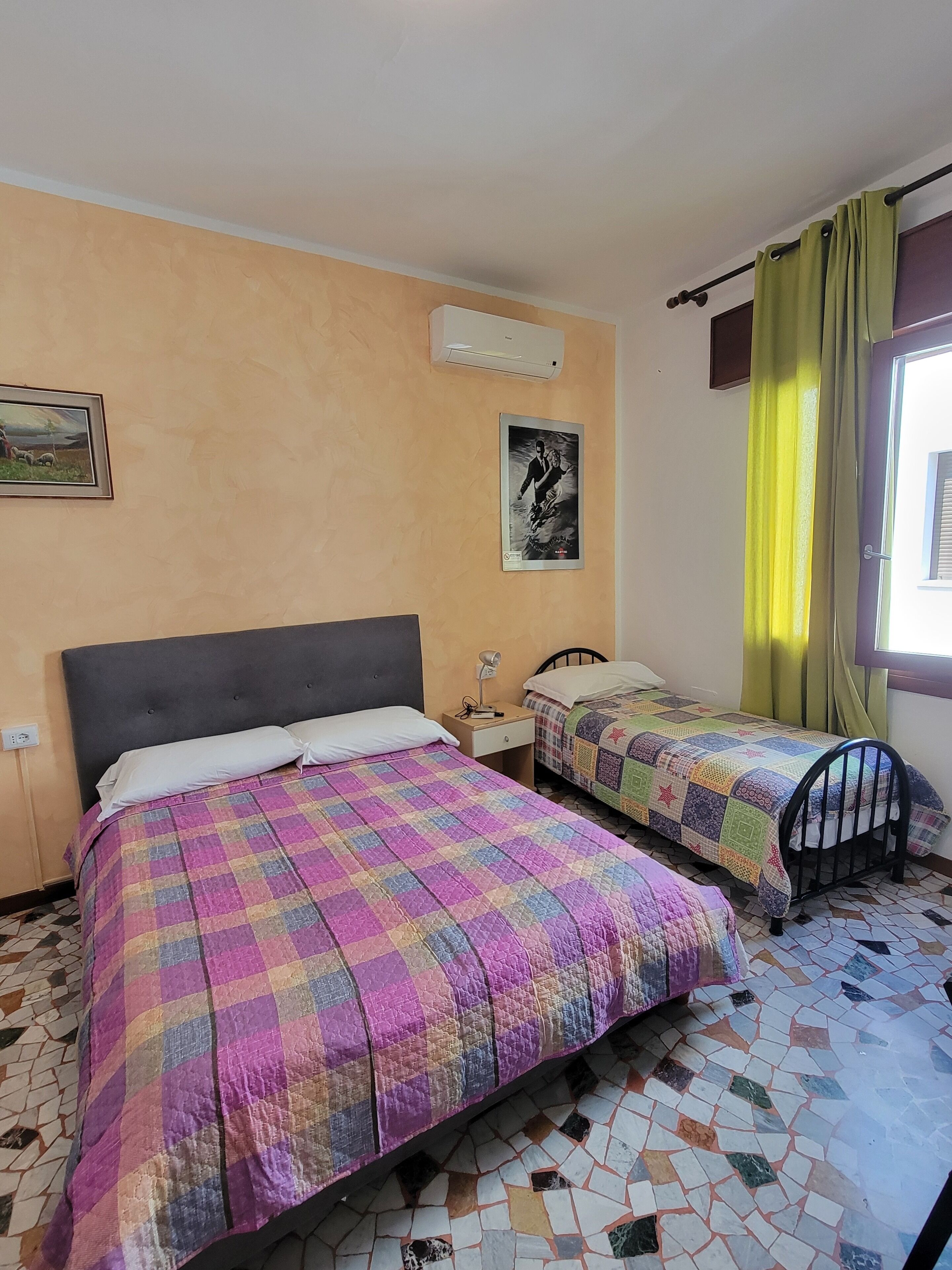 A room with two beds, a wall-mounted air conditioner, a framed picture, and a window with curtains.