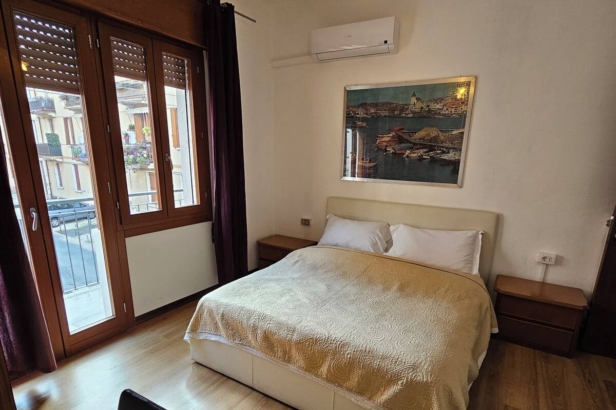 Une chambre d’hôtel avec un lit, un bureau, une chaise, un lustre, un tableau et une vue sur les bâtiments par la fenêtre.