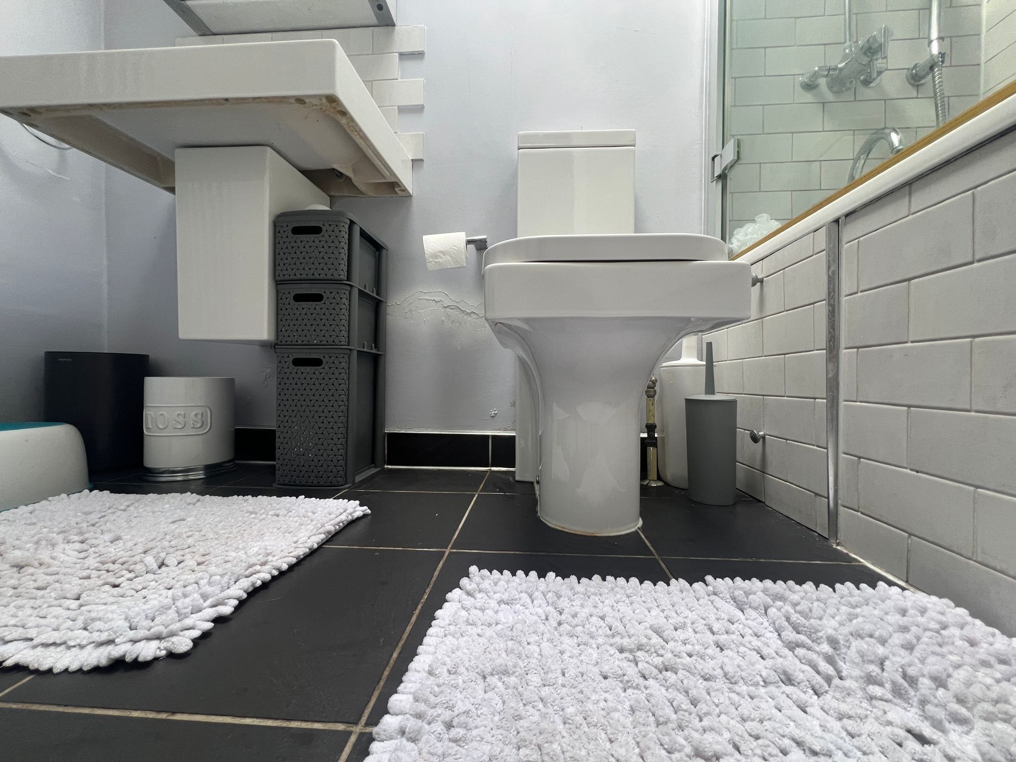 A modern bathroom with a toilet, sink, and shower. There are white rugs on the floor and storage bins against the wall.