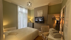 Una habitación de hotel con cama, una silla, un escritorio con lámpara, televisor y ventana con cortinas.