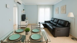 A living room with a black leather sofa, a glass dining table with green placemats, and a TV on a white cabinet. - Oliva Nova Club Sevilla VI Equestrian Center . Playa 400 Metros (OLIVA)