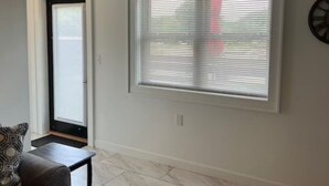 A room with a door, a window with blinds, a patterned pillow on a couch, and a small table.