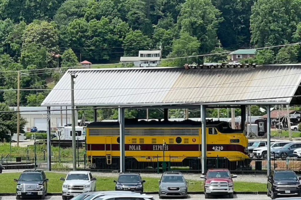 Ein gelb-roter Zug mit der Nummer 4210 fährt durch einen Bahnhof, umgeben von geparkten Autos und Grünflächen.