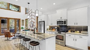 Una cocina moderna con armarios blancos, una isla central con taburetes, electrodomésticos de acero inoxidable y un ventanal con vistas a un área boscosa.