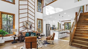 Amplia sala de estar con cocina moderna, una escalera de madera y ventanas grandes con vistas al exterior.