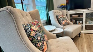 A living room with two beige armchairs, a round coffee table, and a TV stand with books and decorative items. - Walkable Cottage, Pet Friendly with Fenced Patio (Southern Pines)