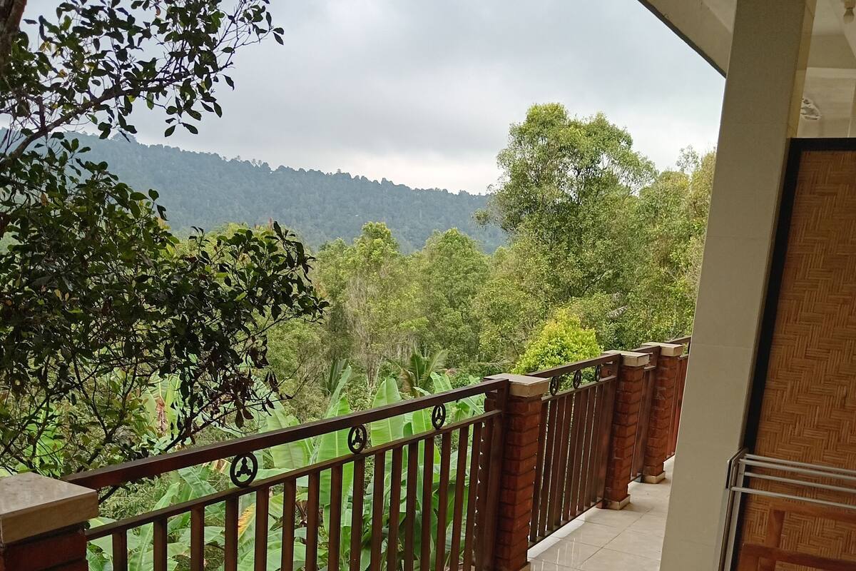 Un balcón con suelo de baldosas, baranda de madera y vista a un paisaje verde y exuberante con árboles y colinas al fondo.