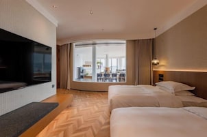Habitación de hotel con un televisor grande, dos camas, piso con estampado de espiga, vista al exterior y un comedor visible a través de la ventana.