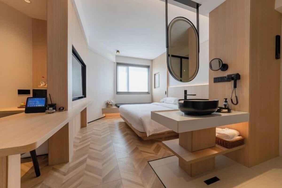 A modern hotel room with a bed, a desk, a large mirror, and a bathroom with a sink and mirror.