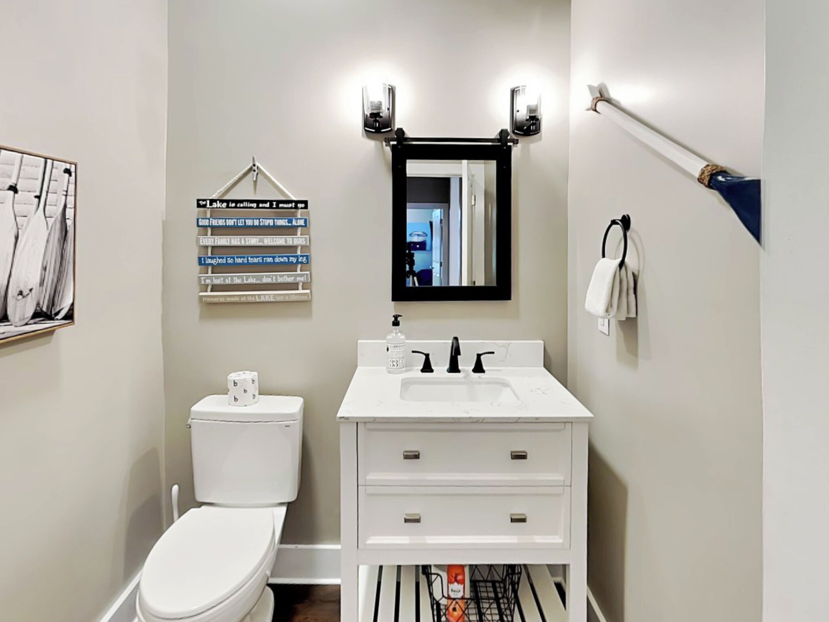A bathroom with a white toilet, a sink with a mirror above it, and a towel rack. There is a sign on the wall with various phrases.