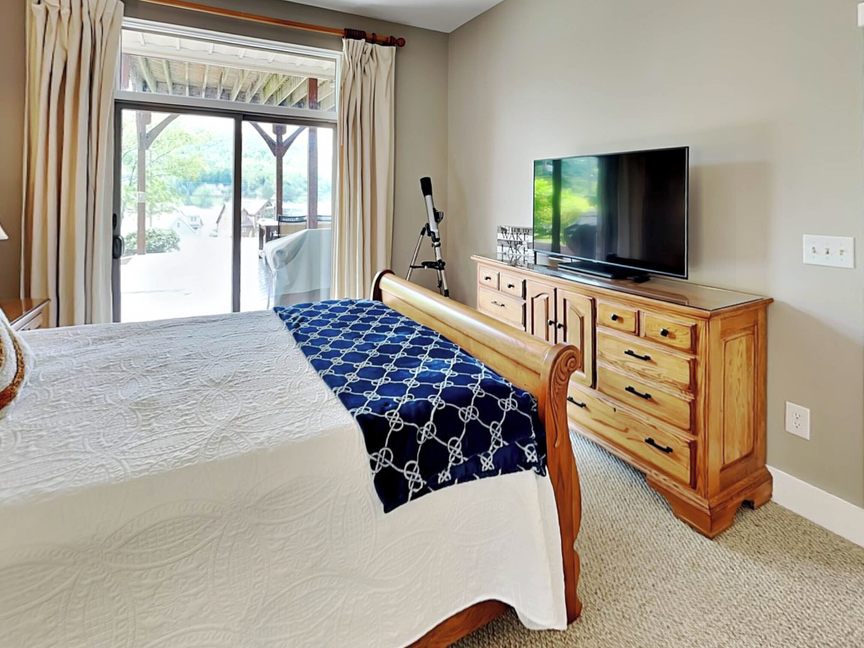 A hotel room with a bed, a wooden dresser, a television, and a sliding glass door leading to an outdoor area.