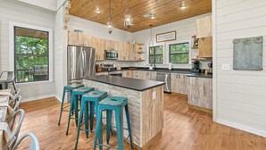 A modern kitchen with wooden cabinets, a large island with bar stools, stainless steel appliances, and a view of the outdoors. - Your Family-Fun HQ w/Hot Tub, Firepit, 2 Gamerooms (Broken Bow)
