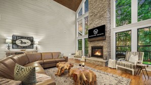 A spacious living room with a stone fireplace, leather sofa, and large windows overlooking a forest. - Your Family-Fun HQ w/Hot Tub, Firepit, 2 Gamerooms (Broken Bow)
