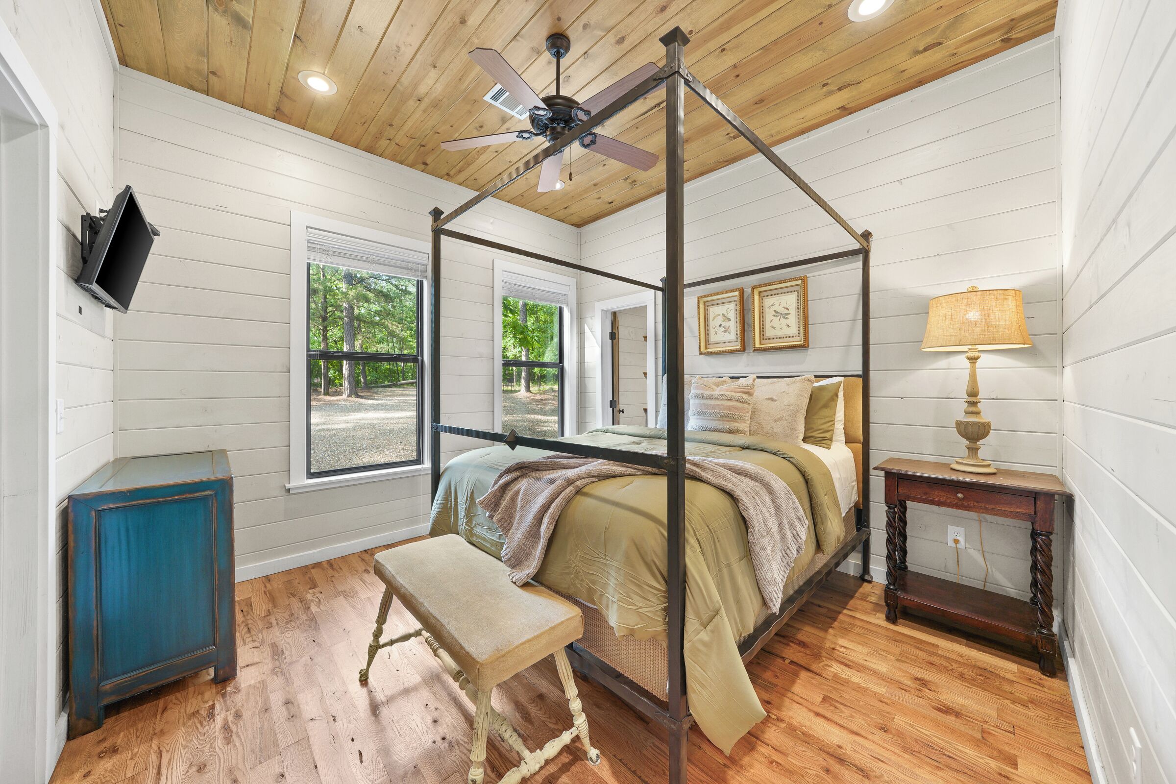 A four-poster bed with a wooden ceiling fan, a bench, a bedside table, and a window with a view of trees.