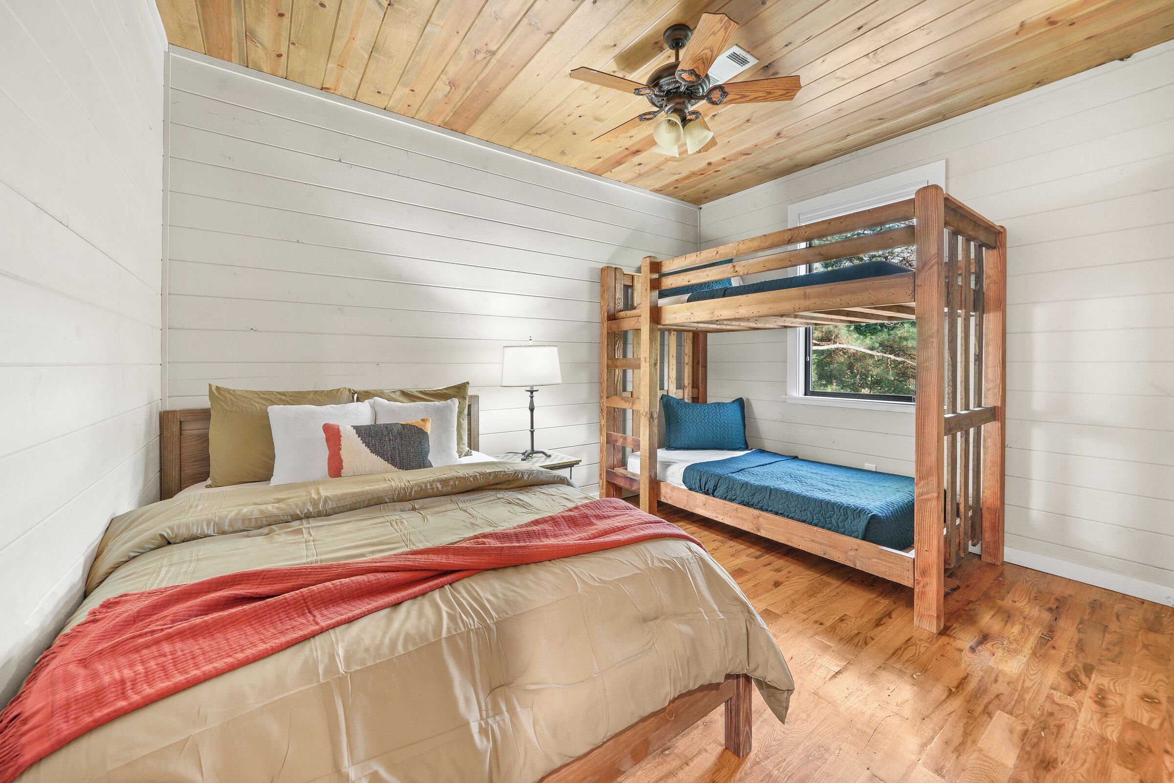 A bedroom with a bunk bed, a single bed, wooden flooring, a ceiling fan, and a window with a view of greenery.