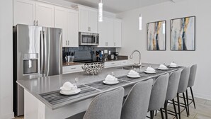 A modern kitchen with stainless steel appliances, a central island with bar stools, and contemporary artwork on the walls. - Luxury 4BR Champions Gate Home w/ Private Pool, BBQ, and Water Park Access! (Davenport)