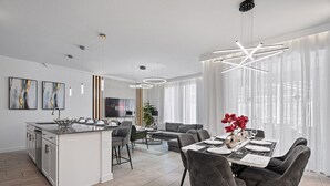 A modern kitchen with a central island, a dining area with a table set for four, and a living area with a sofa and a TV. - Luxury 4BR Champions Gate Home w/ Private Pool, BBQ, and Water Park Access! (Davenport)