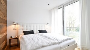 Ein modernes Schlafzimmer mit einem großen Bett, zwei Nachttischlampen, einem Nachttisch und Blick ins Freie durch große Fenster.