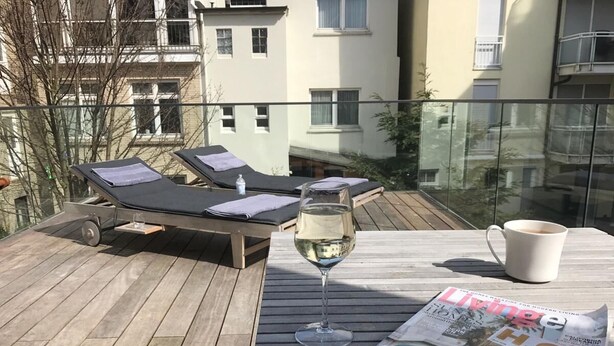 Eine Holzterrasse mit zwei Liegestühlen, einem Glas Weißwein, einer Zeitschrift und einer Kaffeetasse.