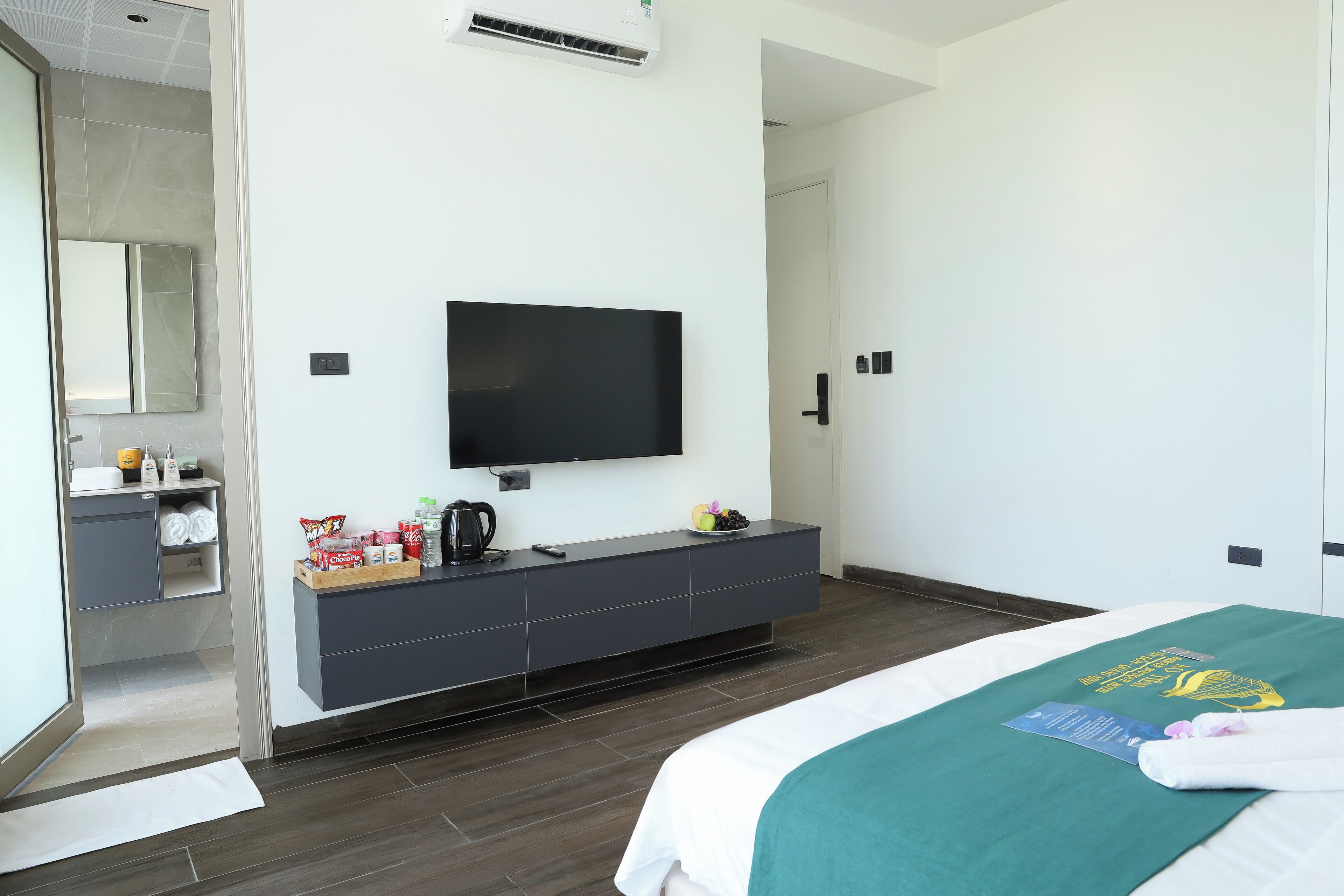 Una moderna camera d'albergo con una TV a schermo piatto, un tavolino grigio con articoli da toilette, un letto con coperta verde e un grande specchio.