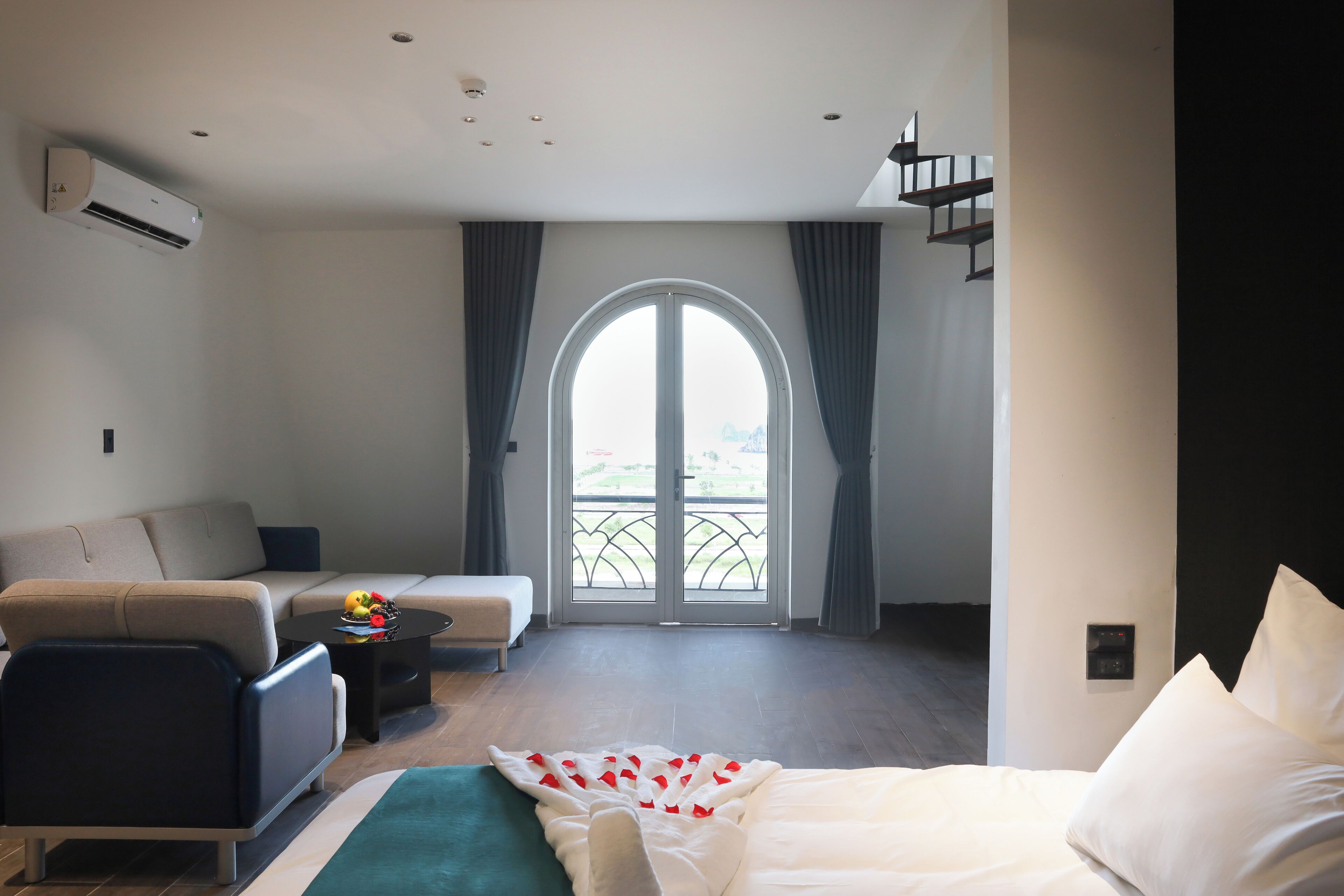 A modern hotel room with a bed, a sofa, a small table with fruit, and a balcony with a view.