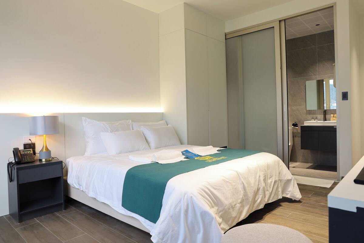 A hotel room with a bed, bedside table, lamp, and a bathroom with a shower and sink.