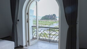 A room with a large arched window, a balcony with a metal railing, and a view of a distant landscape with greenery and a mountain. - Ao Tien Harbour Boutique Hotel (Van Don)