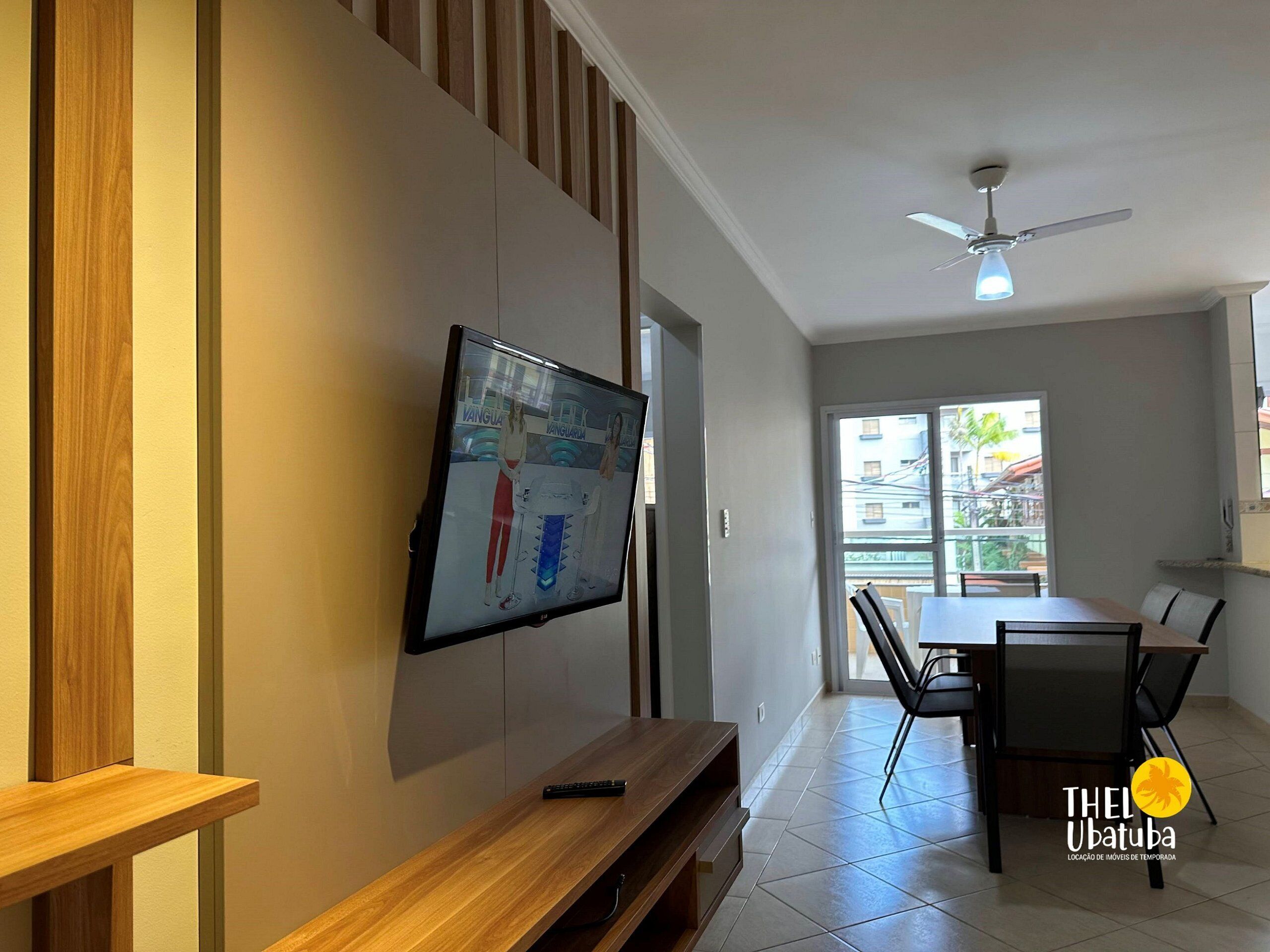 Een moderne woonkamer met een flatscreen televisie aan de muur, een houten bank, een eettafel met stoelen en een plafondventilator.