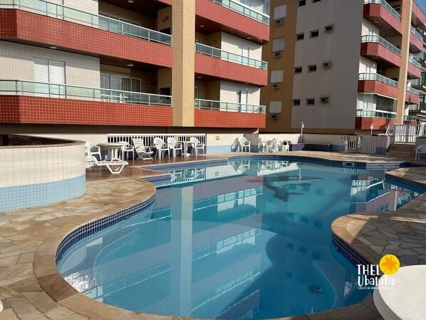An indoor swimming pool with tiled walls and a curved design, surrounded by lounge chairs and a multi-story building with balconies.