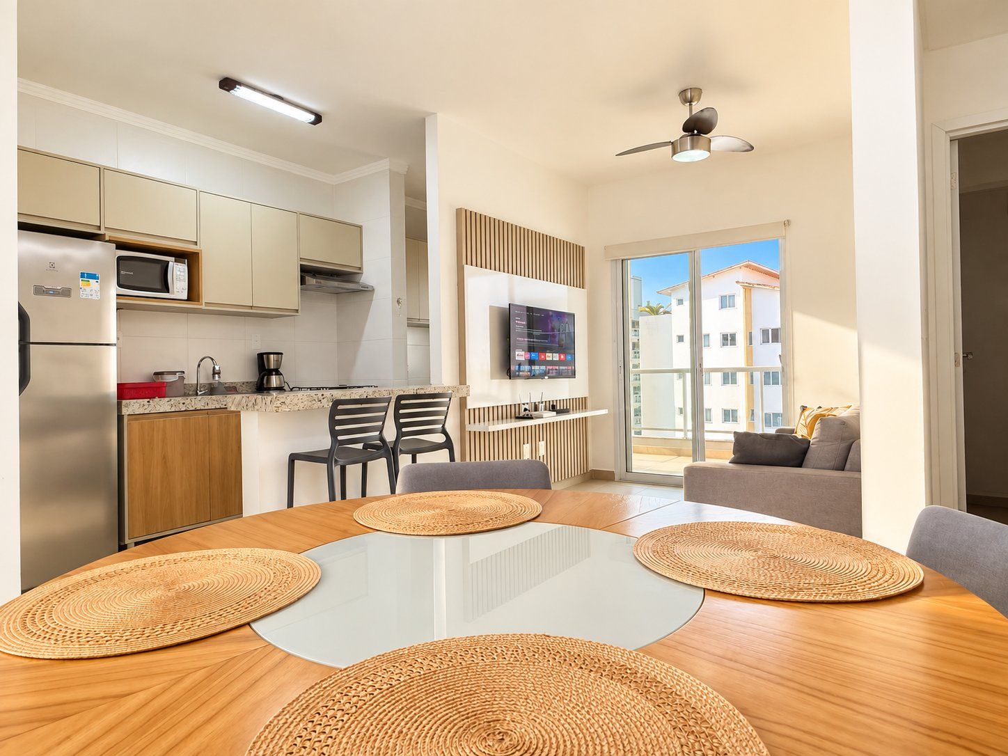 A modern kitchen with a dining area, a TV mounted on the wall, and a balcony with a view of buildings.