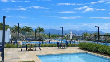 A pool area with chairs and tables, surrounded by a fence and palm trees, with mountains in the background.