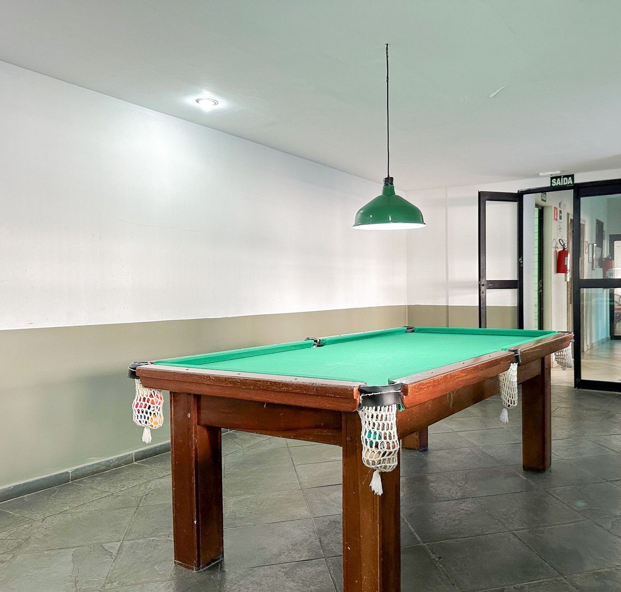 Uma mesa de bilhar com superfície em feltro verde, cercada por uma parede de cor clara e um chão escuro.