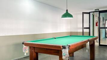 Uma mesa de bilhar com superfície em feltro verde, cercada por uma parede de cor clara e um chão escuro.