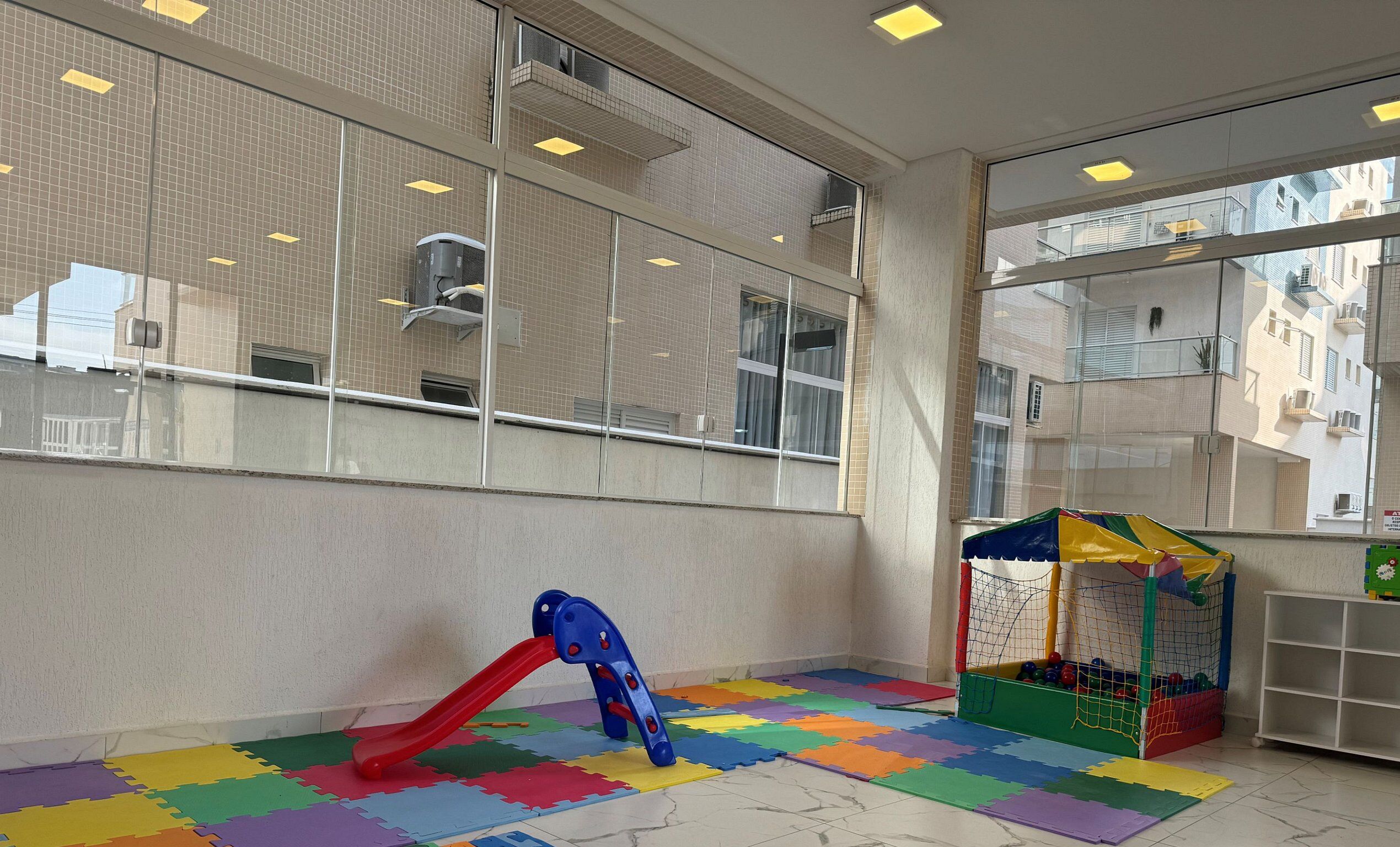 Zona infantil con tobogán, casita de juegos y un ventanal con vistas a edificios.