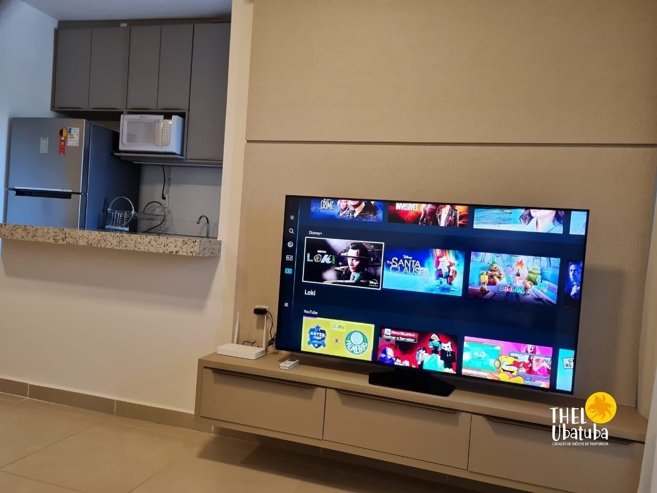 Un televisor de pantalla plana montado en la pared, con una zona de cocina arriba que incluye un microondas y un refrigerador.