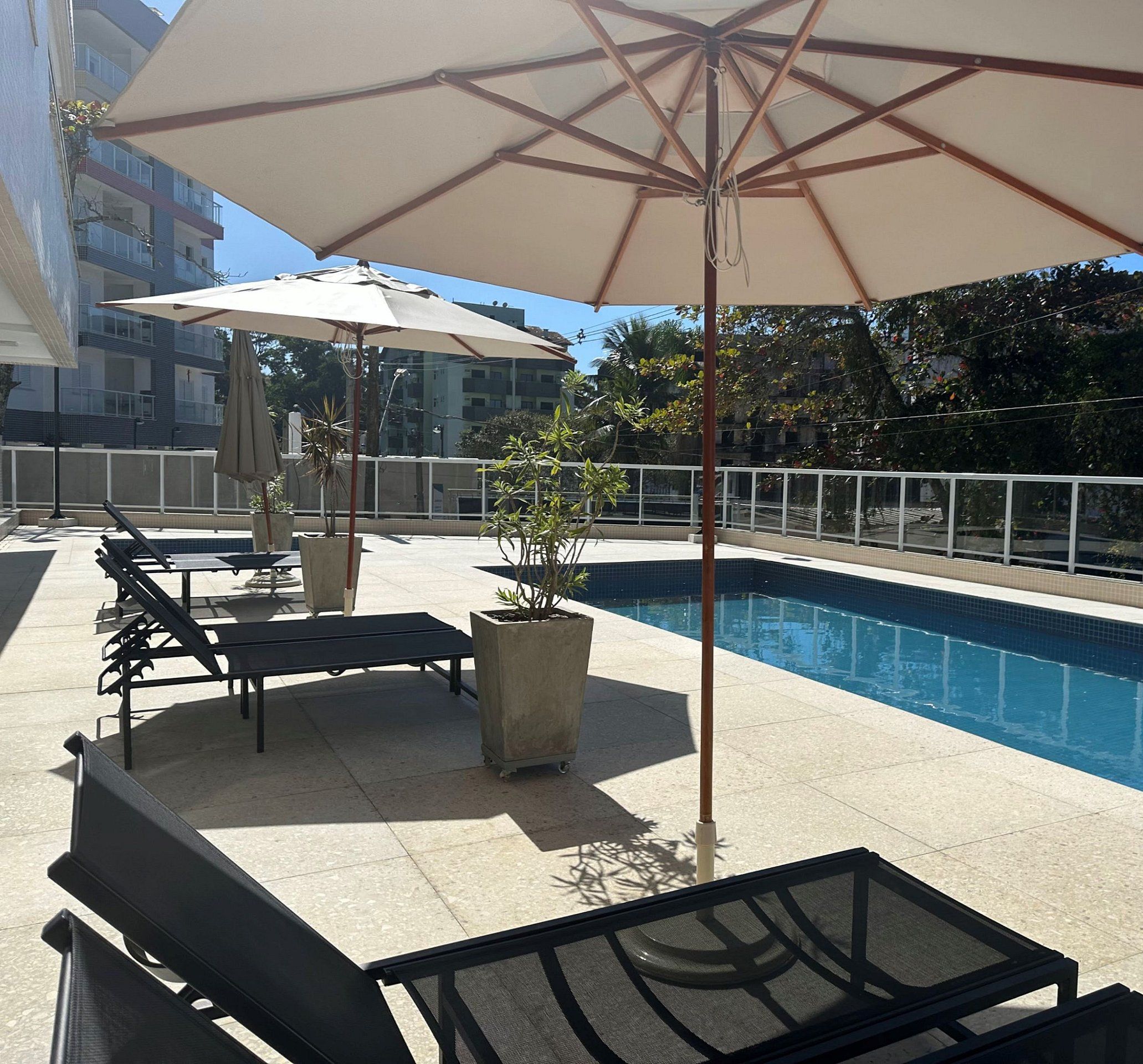 A swimming pool with lounge chairs and umbrellas, surrounded by a white fence and buildings in the background.
